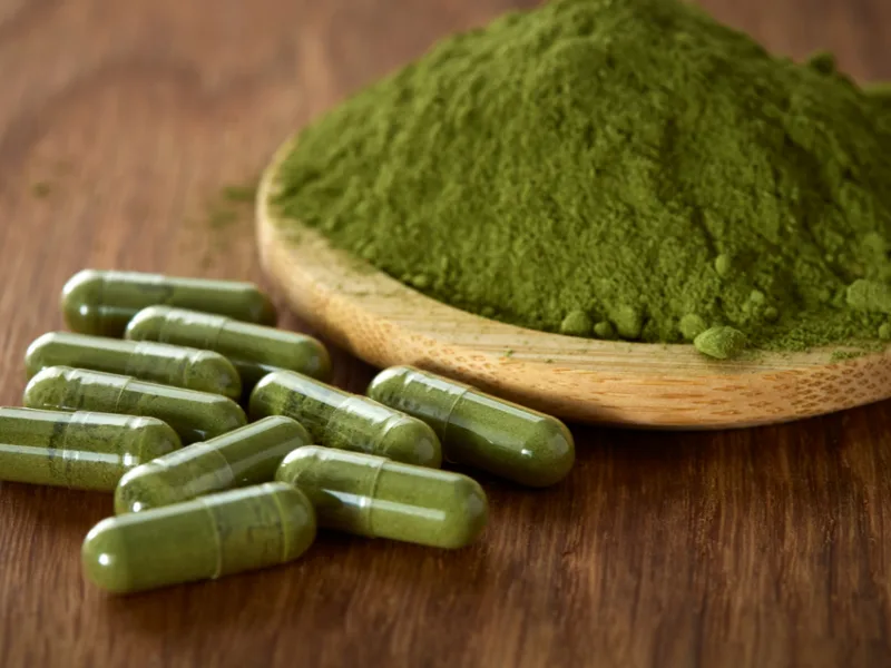 Organic moringa powder in a wooden spoon alongside plant-based pullulan capsules filled with moringa 