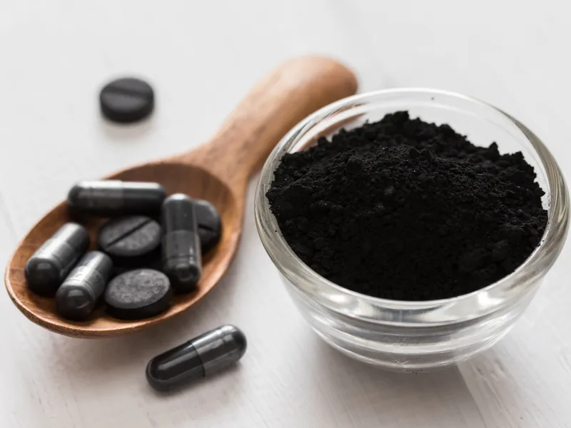 Activated charcoal powder in a glass bowl with charcoal capsules and tablets on a wooden spoon against a clean light background